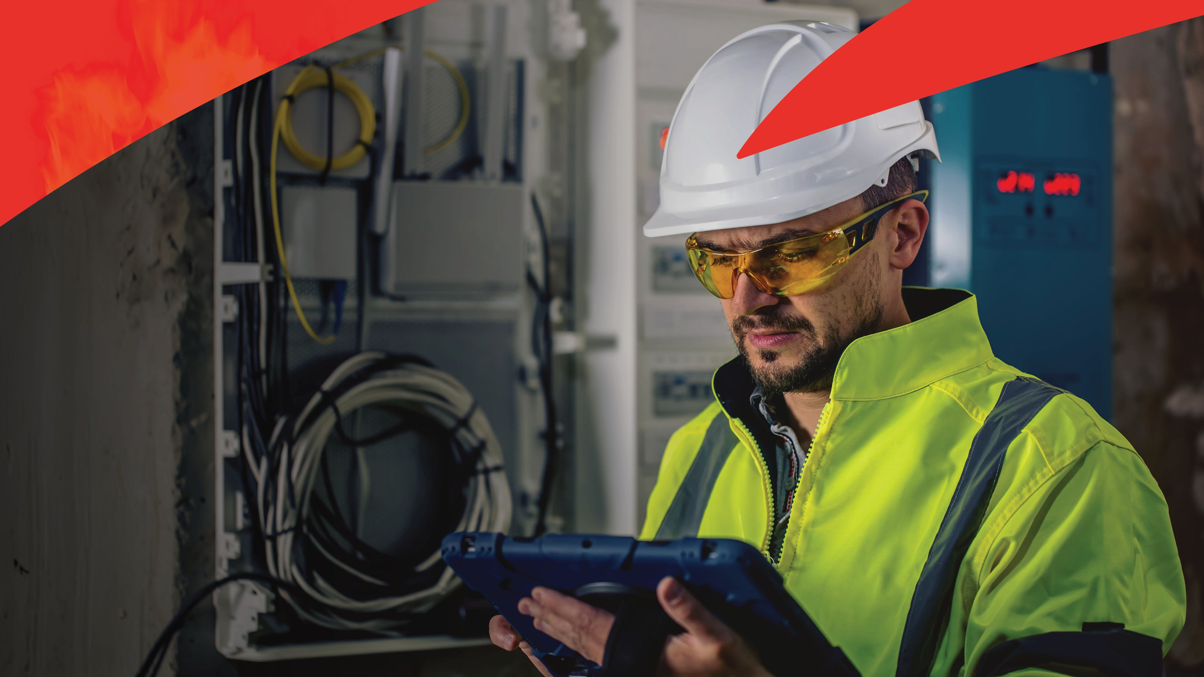 Worker in a high-visibility jacket & hat examines a tablet in control room with cables & equipment.